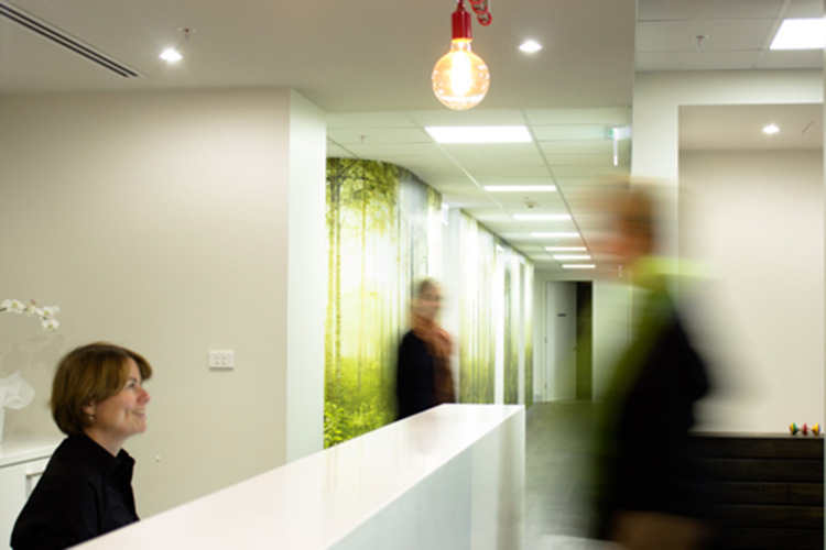 A reception staff member sits behind a front desk in a radiology clinic while people move through the reception area.