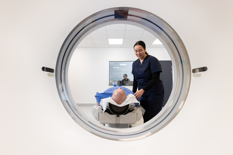 Man lying on bed of CT machine with female radiographer explaining scan to him and another female radiographer in the background setting the machine up