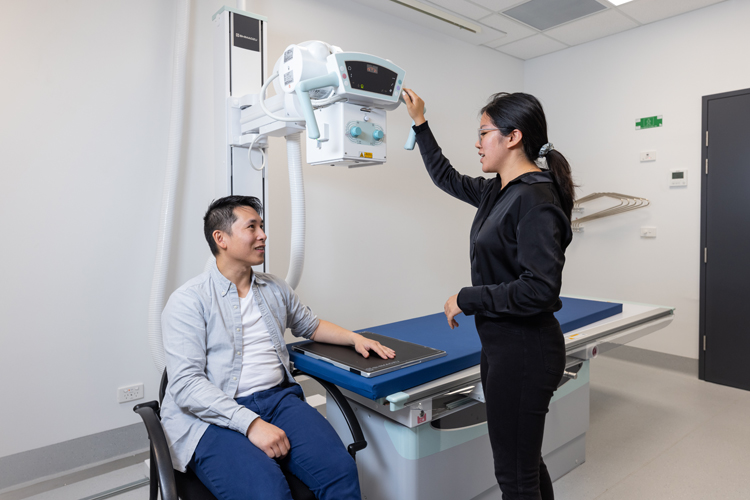 An X‑ray technician positions an overhead X‑ray unit above a seated patient in an imaging room.