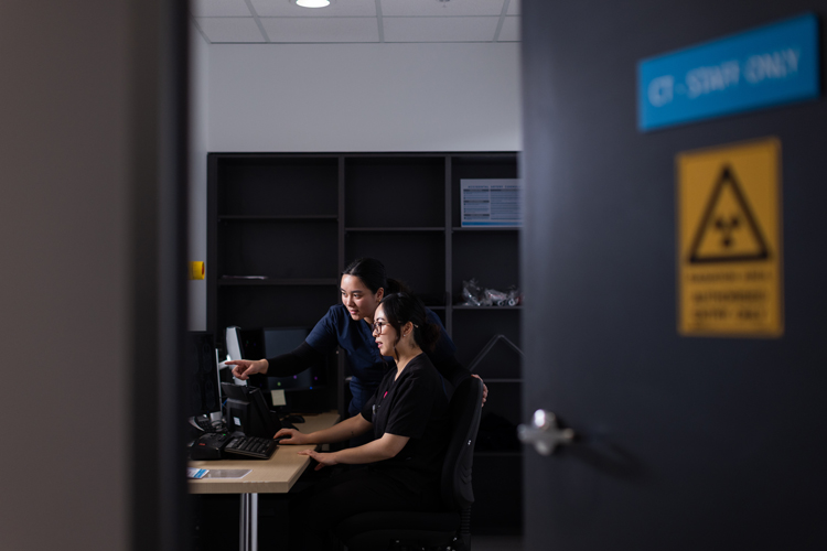 Two female radiographers reviewing images as the CT machine captures them.