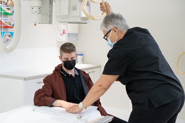 An X‑ray technician positions a patient’s hand on an imaging plate while adjusting overhead equipment in a clinical imaging room.
